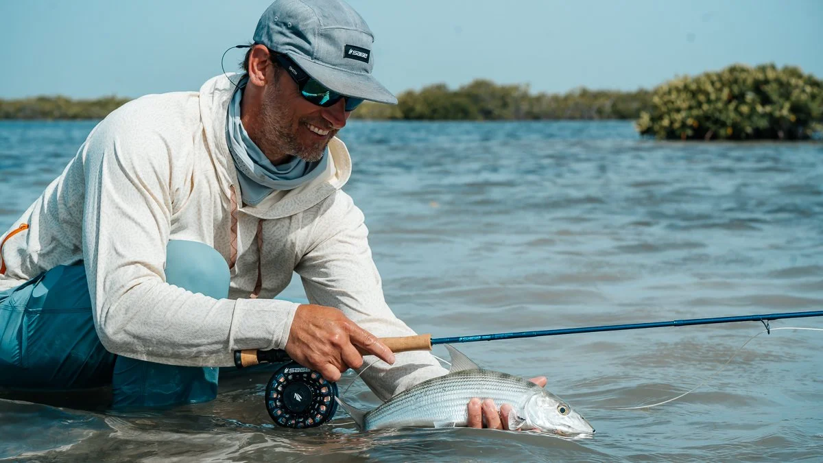 Man in outdoor fishing gear holding a fish with a fishing rod near water with trees in the background.