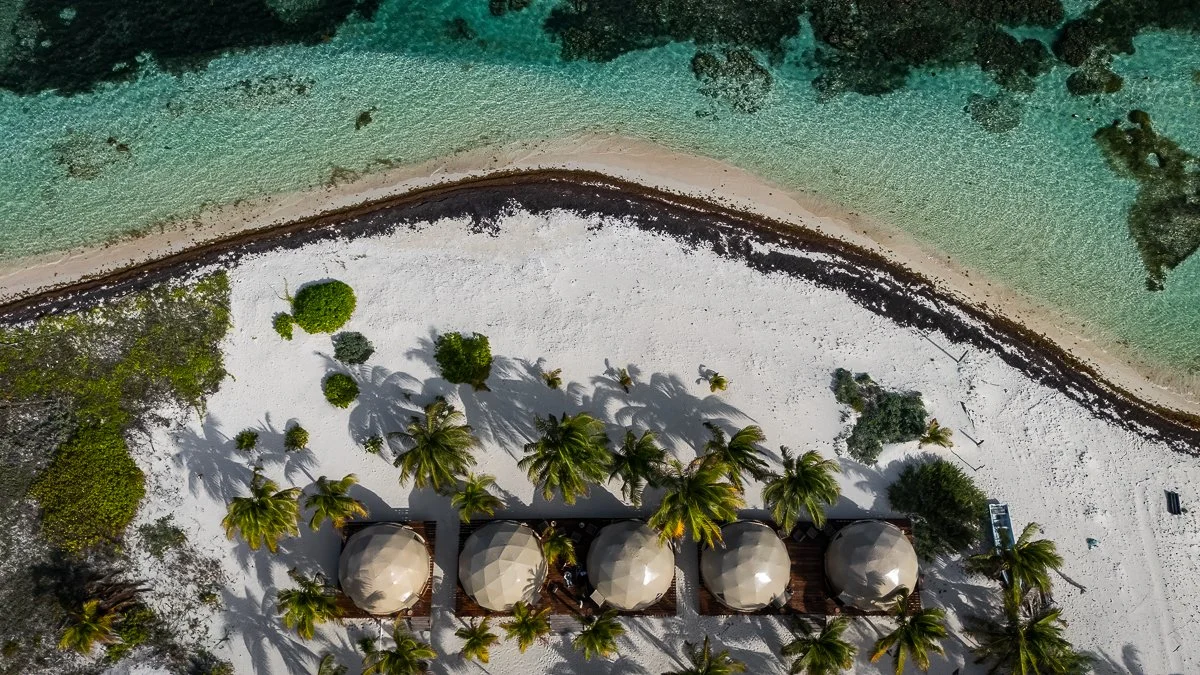 Aerial view of a beach with white sand, turquoise water, and palm trees lining a row of tents or huts.