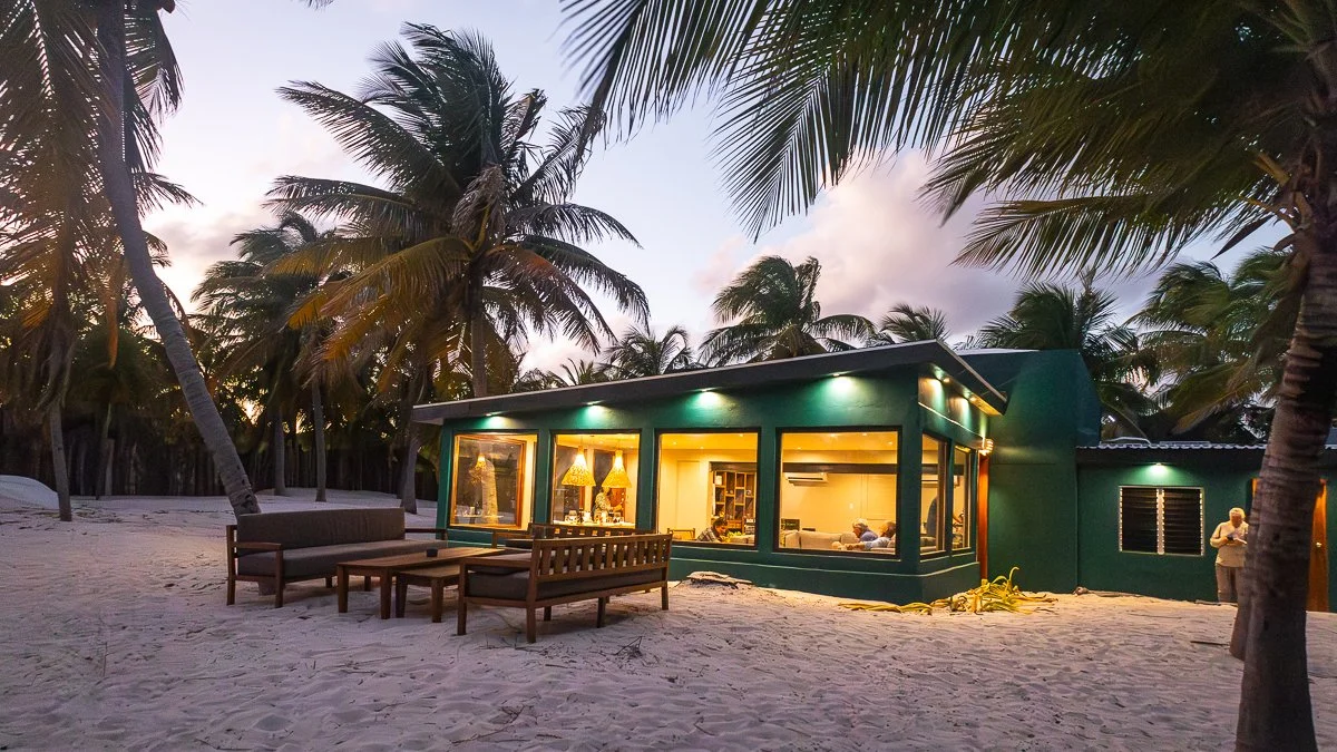Beachside house with large windows, surrounded by palm trees, illuminated indoor lighting, sandy ground, outdoor seating, evening sky