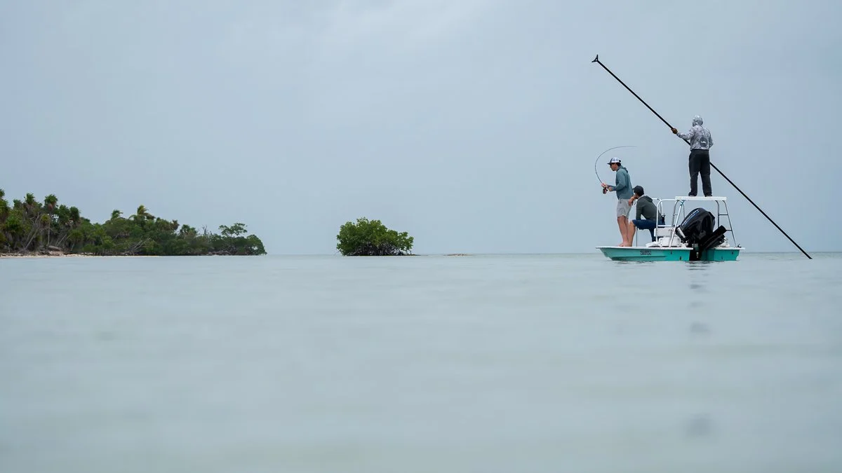 Three people fishing from a small boat on calm water near a small island with trees, overcast sky.
