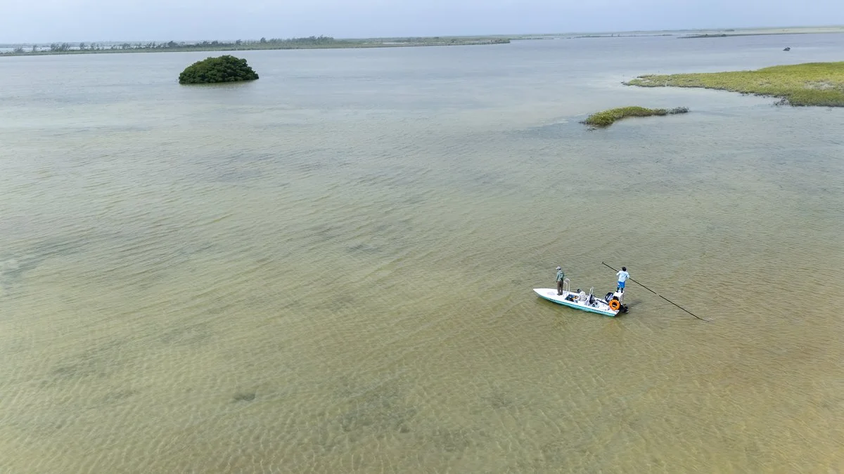 Two people on a boat in shallow water, one fishing with a pole and the other standing near the boat, with small patches of grass and a distant tree island in the water.