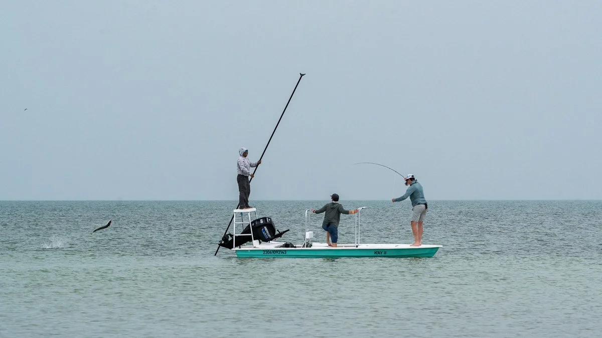 Three people fishing on a boat in the water, with one person standing on a raised platform casting a fishing line, and another person also casting a line, while a third person stands in the middle of the boat.