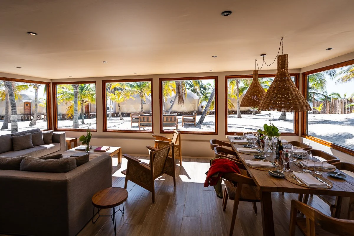 Interior of a beachside living and dining area with large windows showing palm trees and sandy outdoors, with a comfy gray sofa, wooden chairs, and a dining table set with glassware and plates