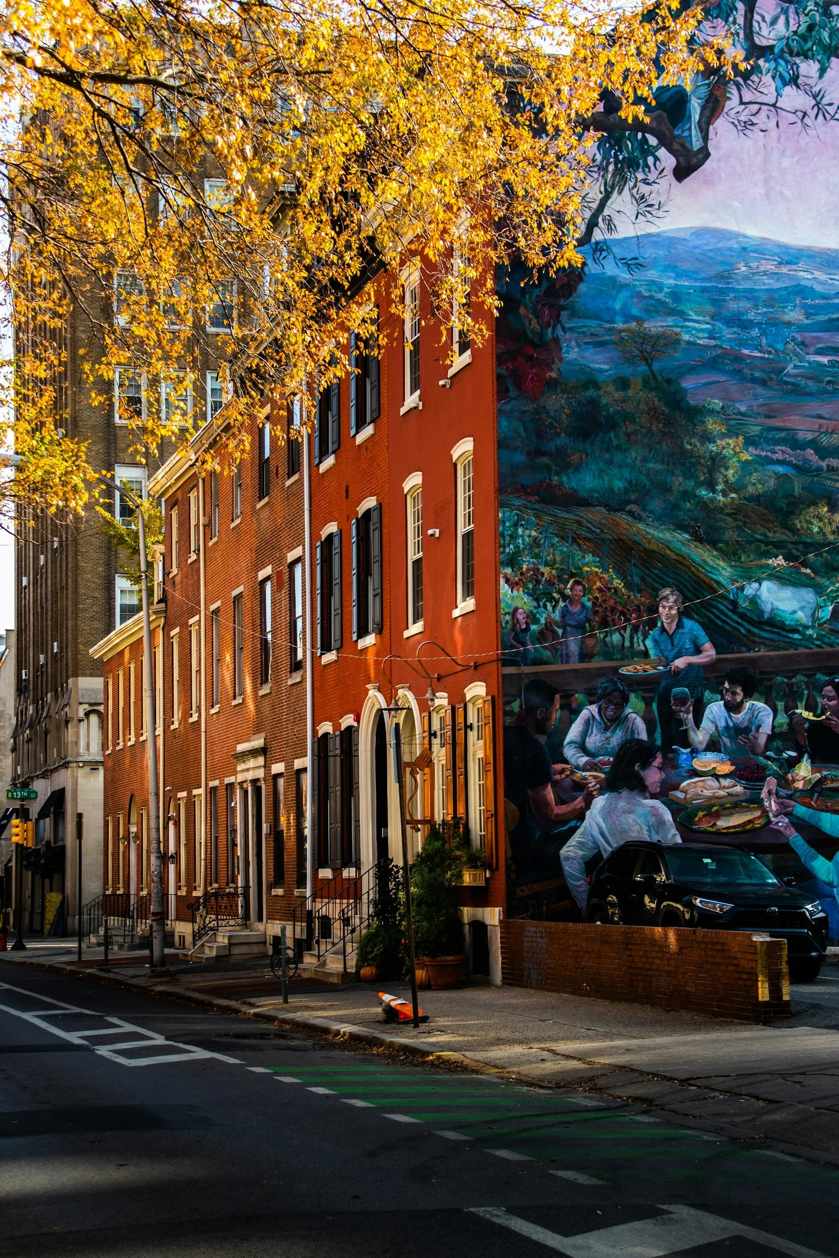 Colorful Philadelphia mural painting on building wall depicting landscape with mountains, trees, and people, with a mustard-yellow tree in the foreground.