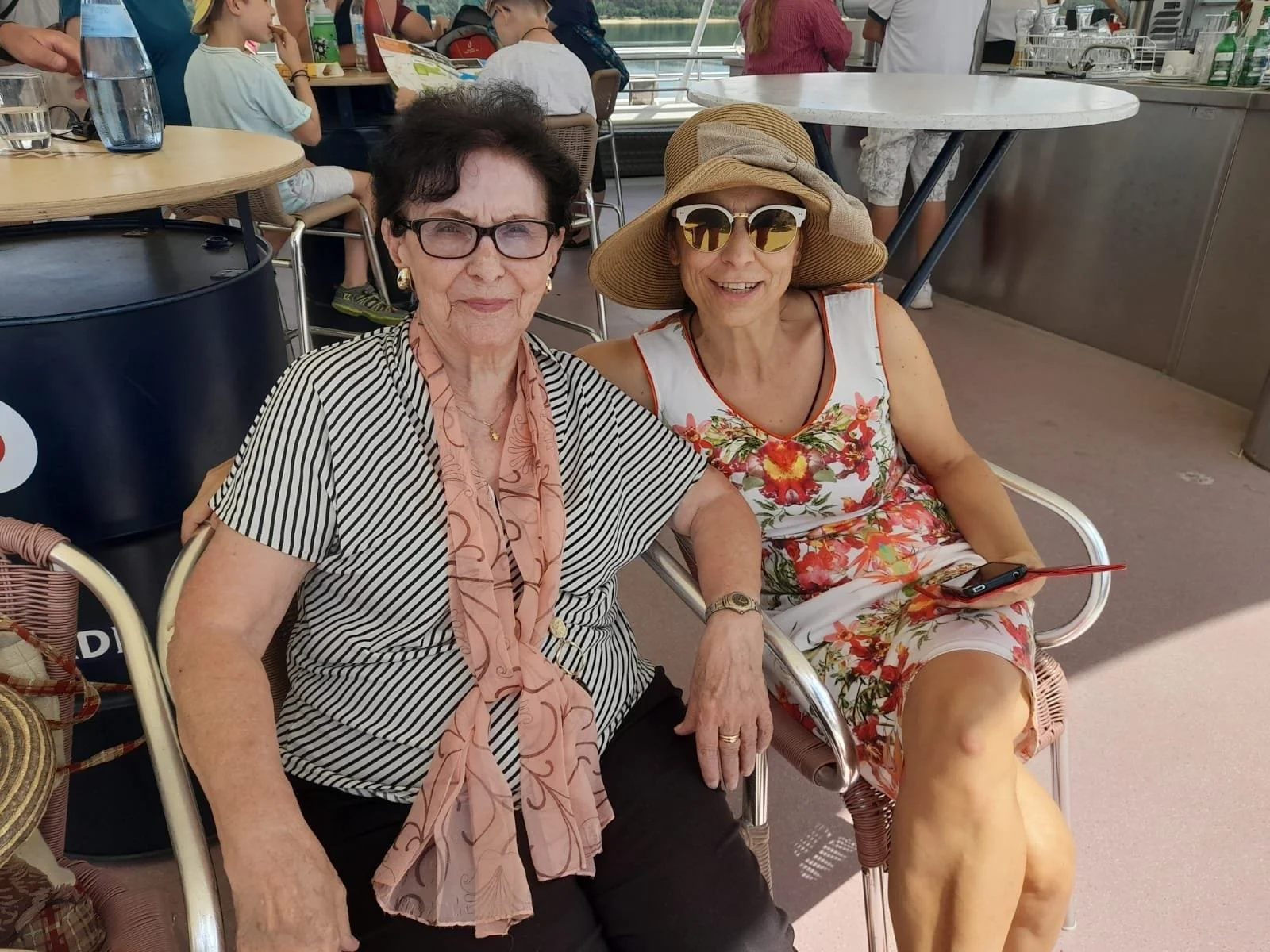 Zwei ältere Frauen sitzen zusammen in einem Café, die eine trägt eine schwarze und weiße gestreifte Bluse, eine goldene Halskette und eine Brille, die andere trägt ein Sommerkleid mit Blumenmuster, eine Sonnenbrille und einen Strohhut, während sie lächeln und relaxen.