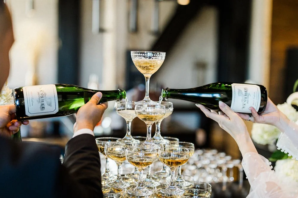 People pouring champagne into a pyramid of glasses at a celebration or wedding event.