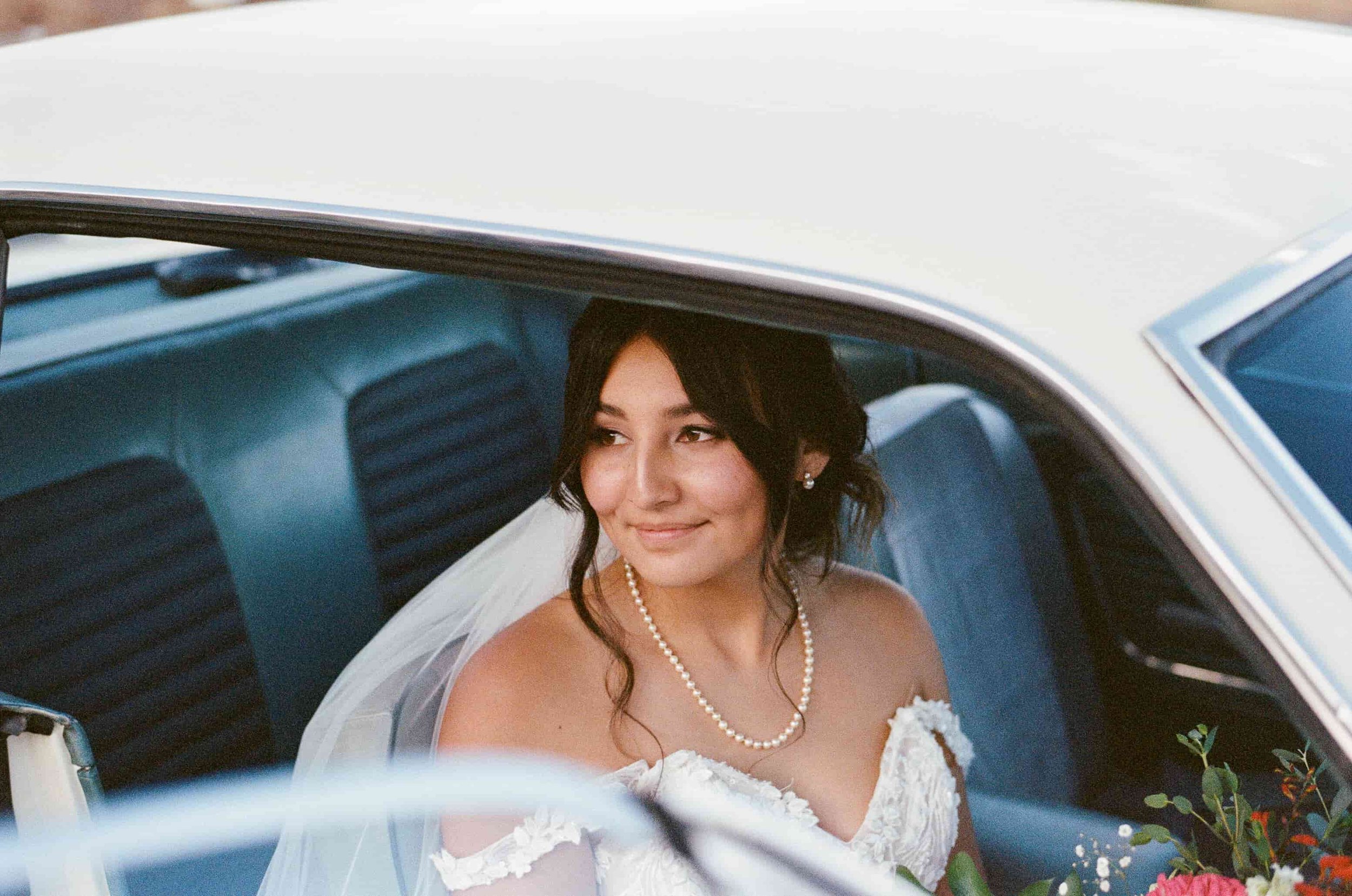 A bride with dark hair and pearl earrings sitting in a vintage car, wearing a white lace wedding dress and veil, smiling and looking out the window, with flowers visible outside the car.