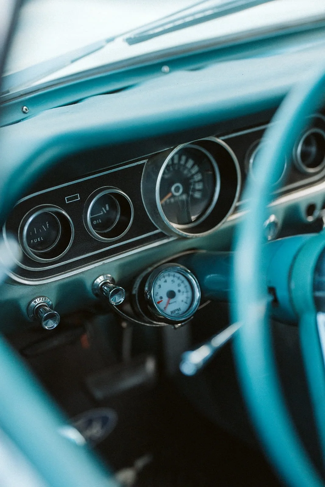 Close-up of vintage car dashboard with speedometer, fuel and oil gauges, and a mechanical tachometer, all in a teal color scheme.