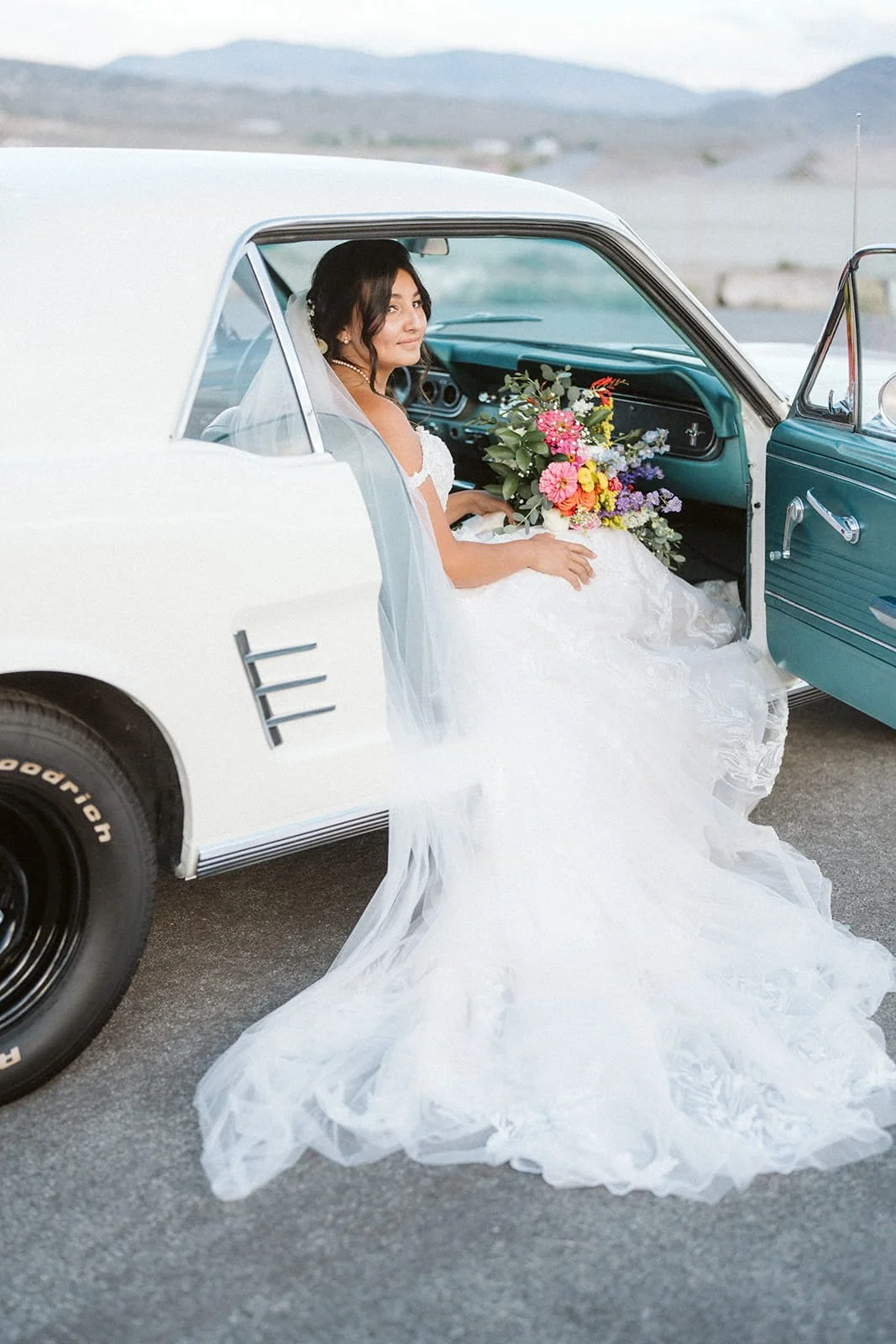 Bride sitting in a vintage white car with the door open, holding a colorful bouquet of flowers, wearing a white wedding gown and veil, in a scenic outdoor setting.