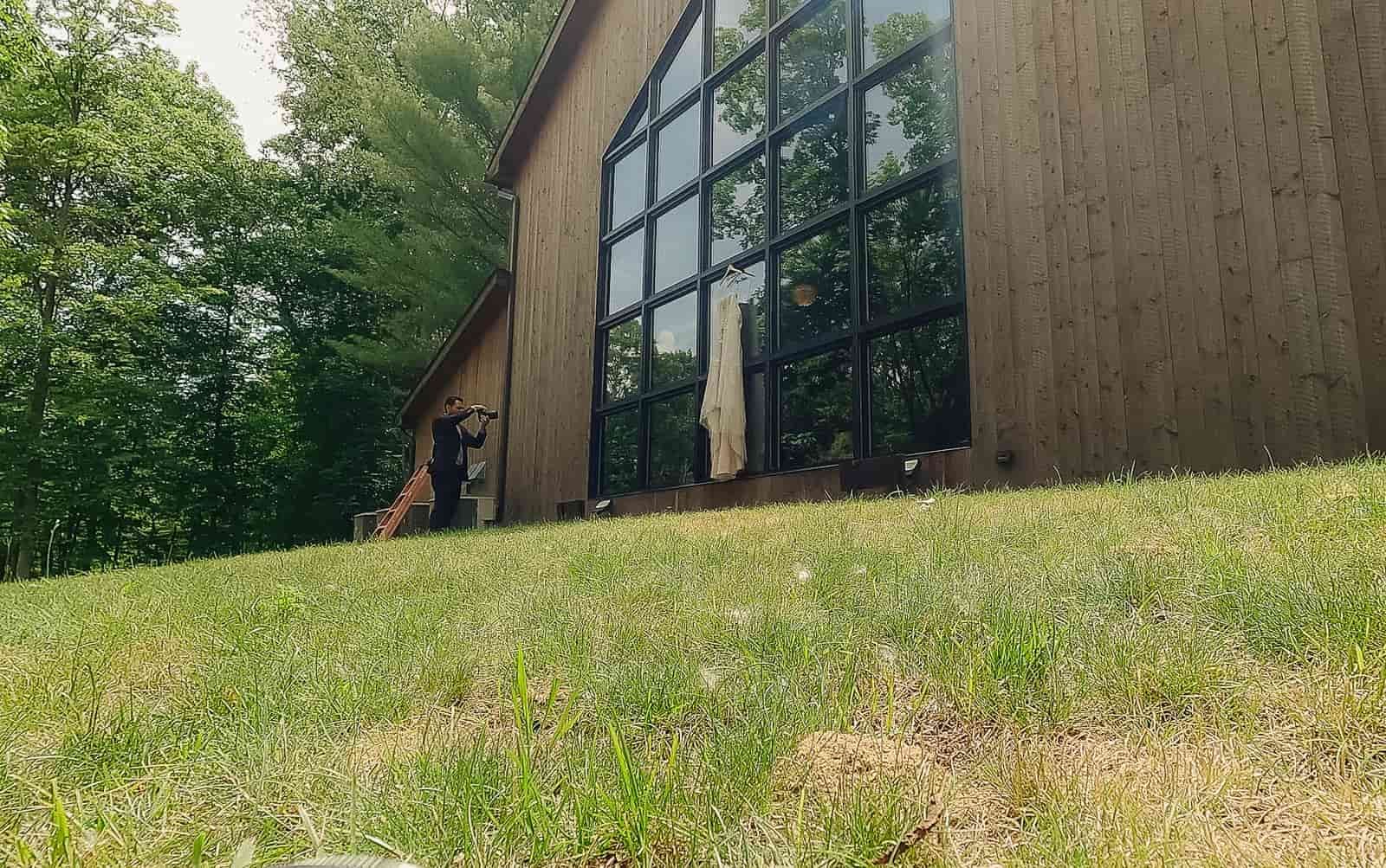 A man in a suit taking a photograph of a dress hanging on a large window of a wooden building surrounded by trees and grass.