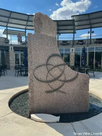 A large abstract sculpture made of pink stone with a metal wire shape on it, set in a circular water fountain surrounded by pavement, with a glass building and outdoor seating in the background.