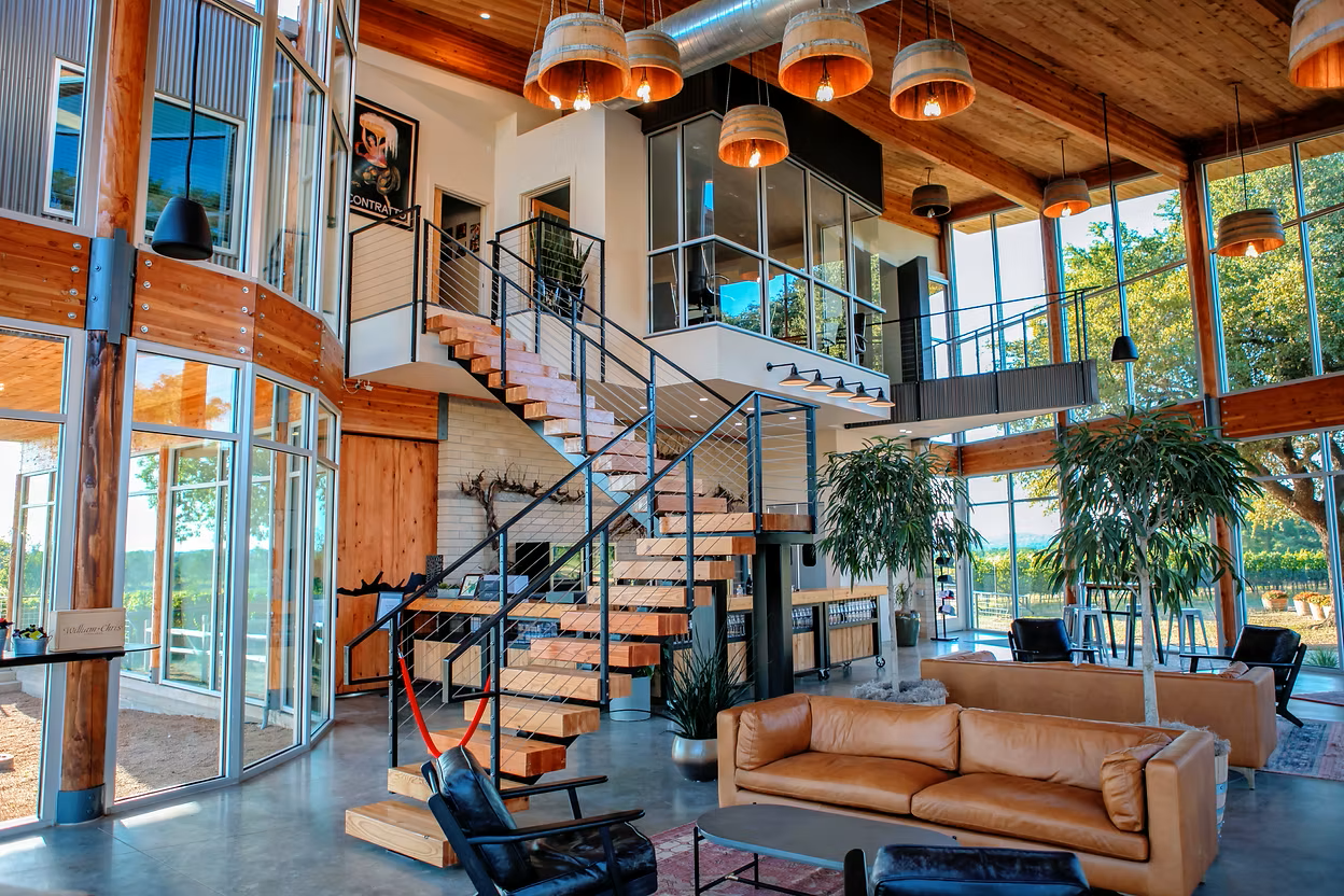Spacious modern interior with large glass windows, wooden ceiling, contemporary furniture, and a staircase with wooden steps and black metal railing.