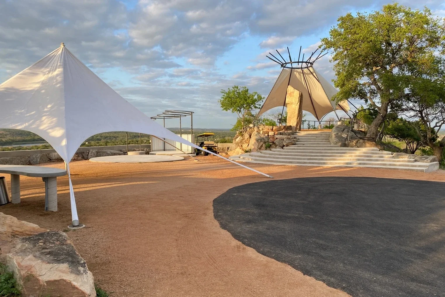Stone steps leading to a shaded canopy structure with a fabric roof and metal poles surrounded by trees under a partly cloudy sky