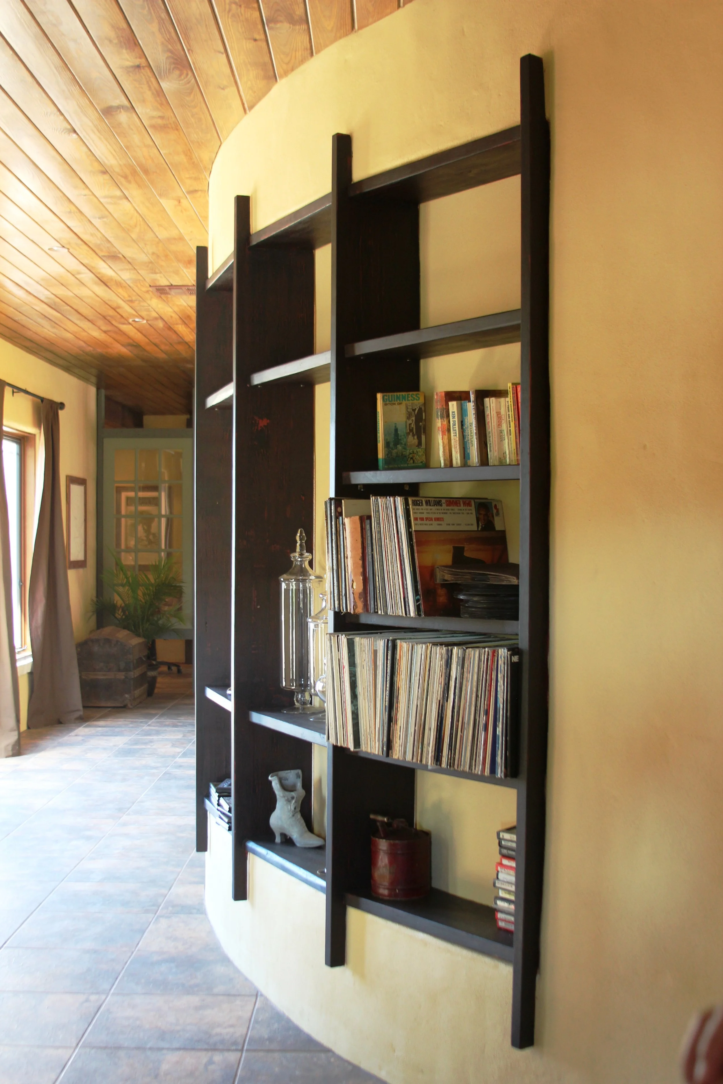 A black bookshelf on a yellow wall holding books, vinyl records, and decorative items in a cozy room with tiled flooring and wooden ceiling.