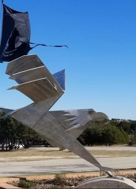 Large metal sculpture of a bird, possibly an eagle, with wings spread and tilted forward, situated outdoors against a clear blue sky.