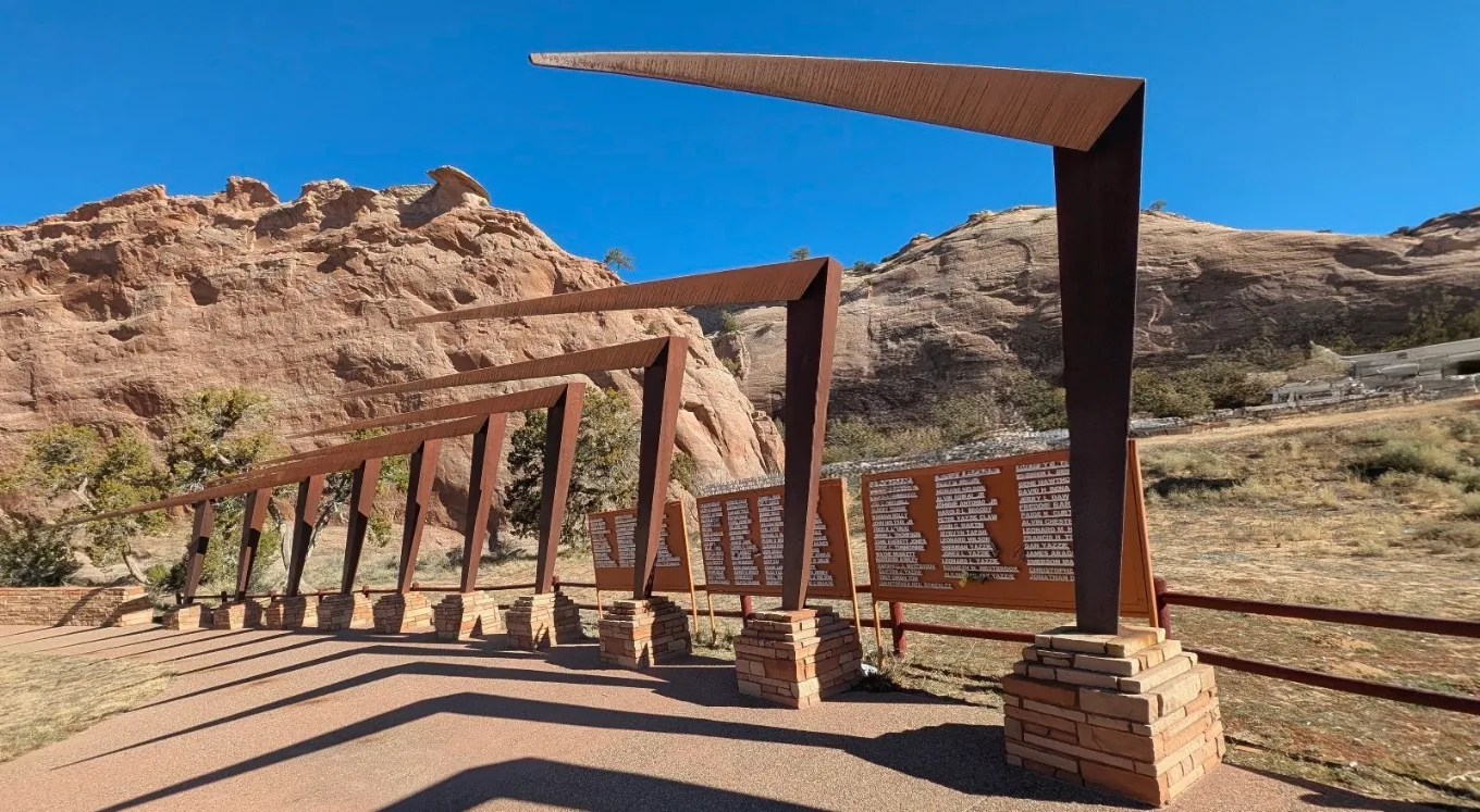 Outdoor memorial with a series of large, rust-colored metal beams on brick bases, arranged in a curve against a rocky desert landscape with a clear blue sky.