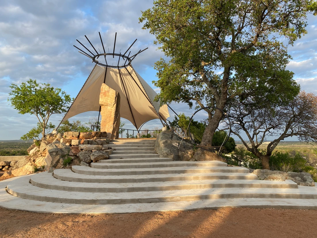 An outdoor stage with a tent-like canopy, stone steps, and trees against a partly cloudy sky.
