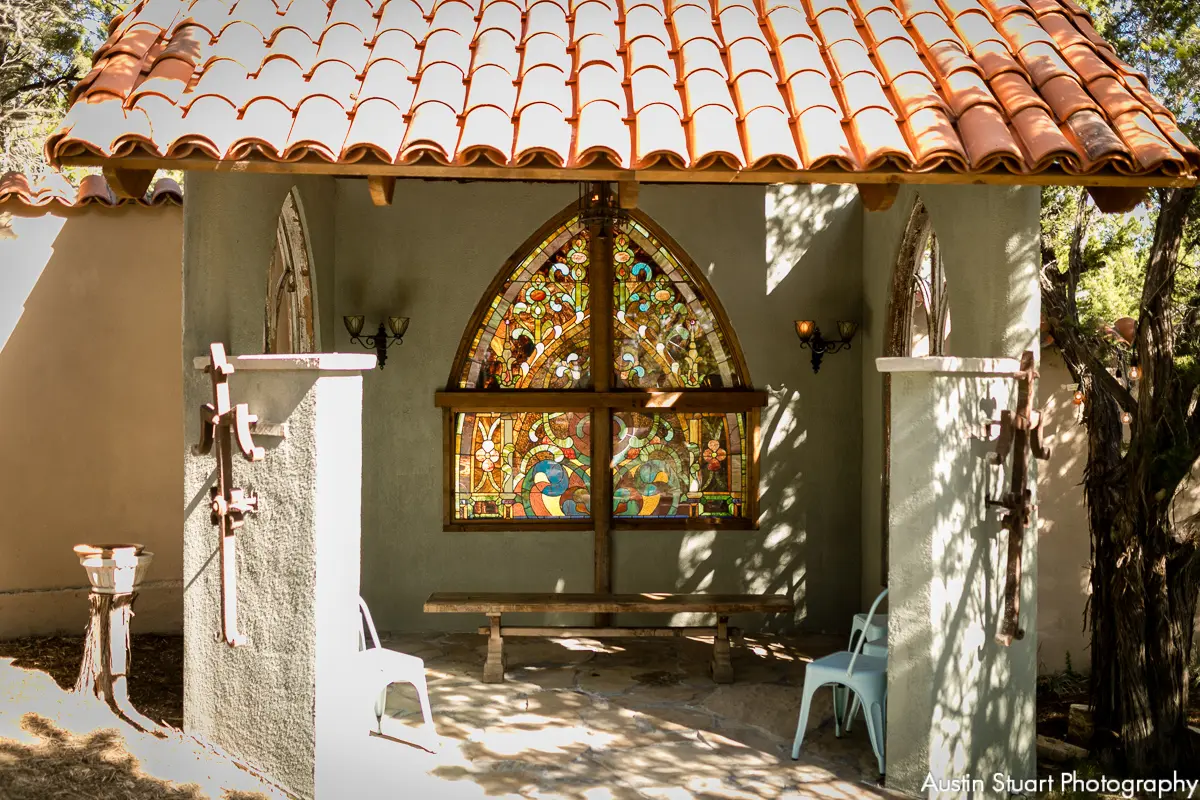 A small chapel with a terracotta tile roof, a large stained glass window, and an open front with two white chairs. Shadows of trees are cast on the structure.