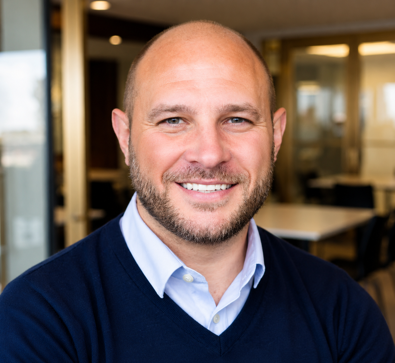 Headshot of a smiling man with a beard and blue eyes, wearing a light blue button-up shirt and a navy sweater, in an office setting.