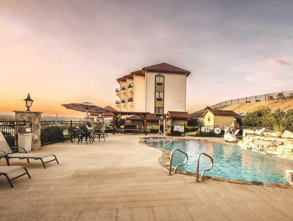 An outdoor swimming pool area at sunset with lounge chairs, tables, umbrellas, and a multi-story building in the background.