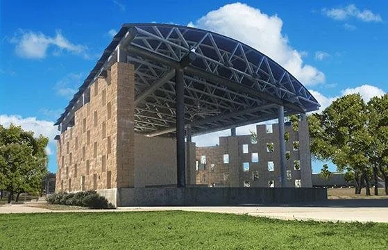 Modern architectural structure with a large metal roof, supported by black pillars, adjacent to a brick wall with window openings, set in a park with green grass and trees, under a partly cloudy sky.