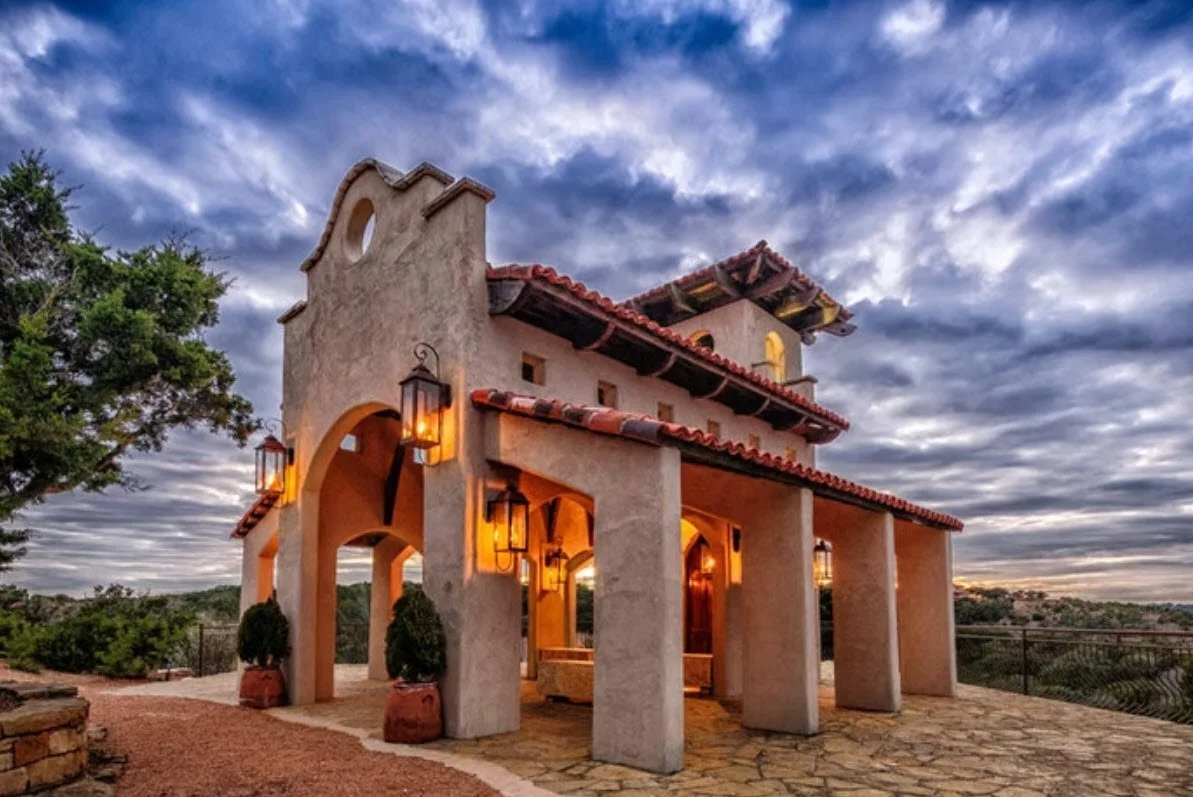 A Spanish-style house with beige stucco walls, red tile roof, arched openings, and lantern-style lights inside and outside. The house is set in a scenic outdoor area with trees and a cloudy sky.