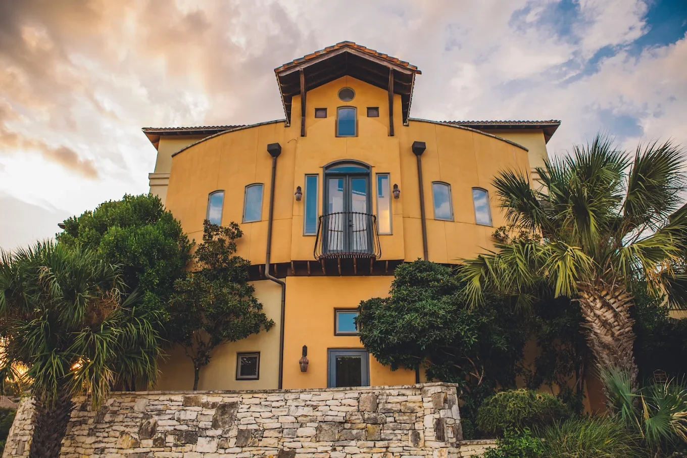 A large yellow house with a stone foundation, surrounded by greenery including palm trees, under a partly cloudy sky during sunset.