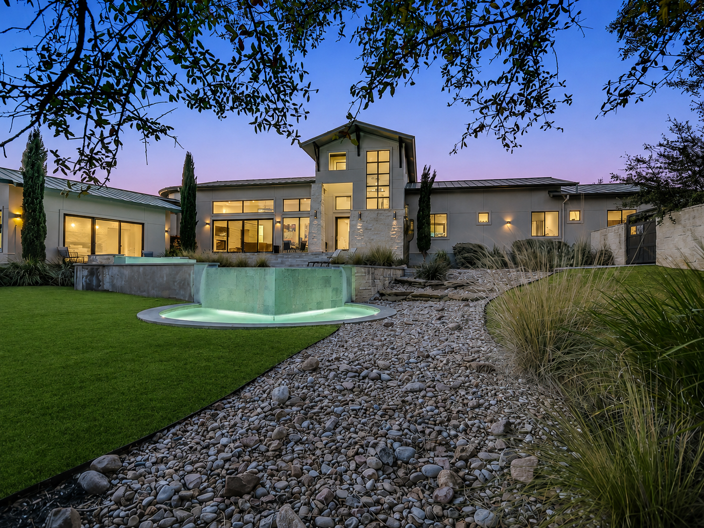 Modern house with large windows, surrounding landscaping, a green lit pool, and stone pathway at dusk.
