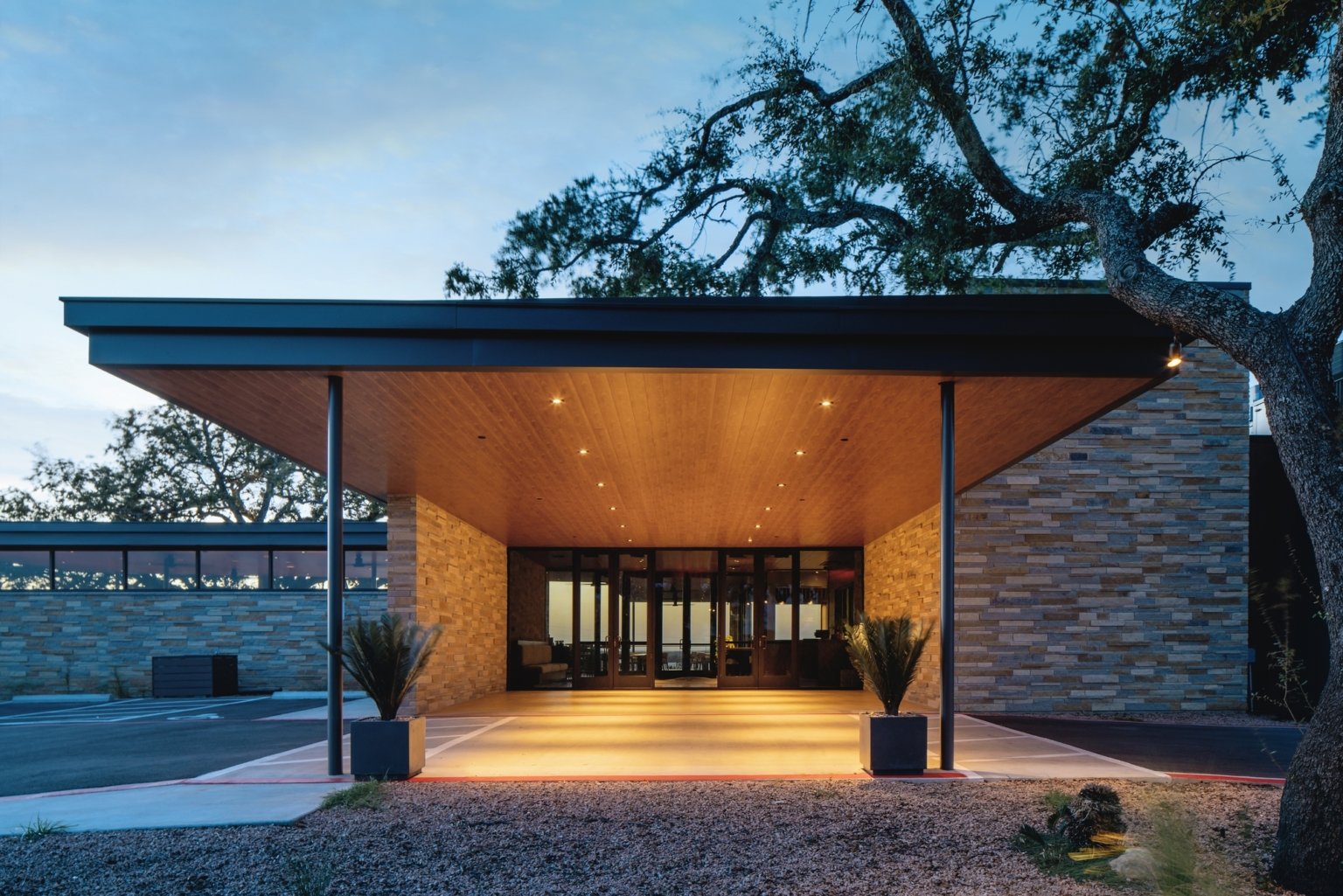 Modern building entrance with a wooden ceiling, stone walls, glass doors, and outdoor lighting, surrounded by trees and parking lot at dusk.