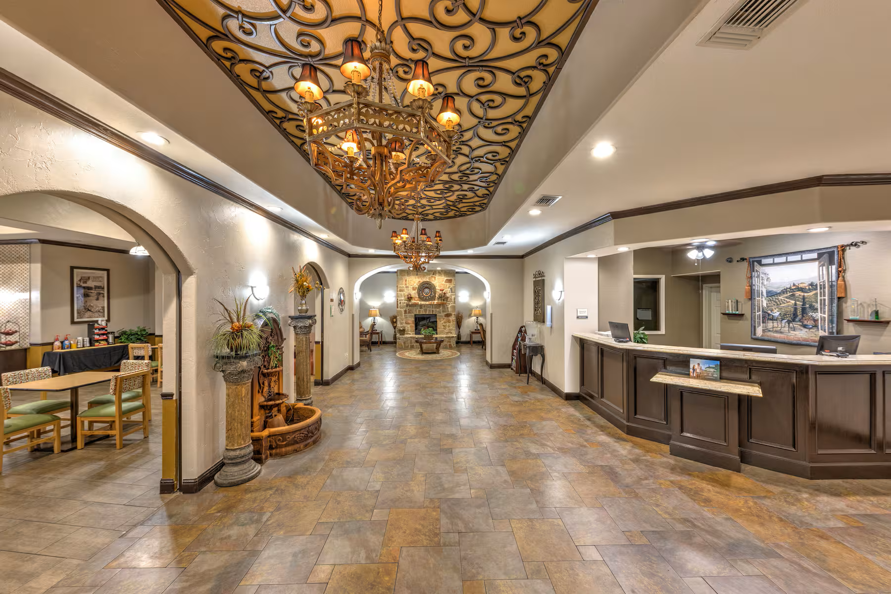 A spacious hotel lobby with a tiled floor, reception desk, a stone fireplace, and elegant chandeliers hanging from a decorative ceiling panel. There are seating areas with chairs, lamps, and decorative plants around the space.