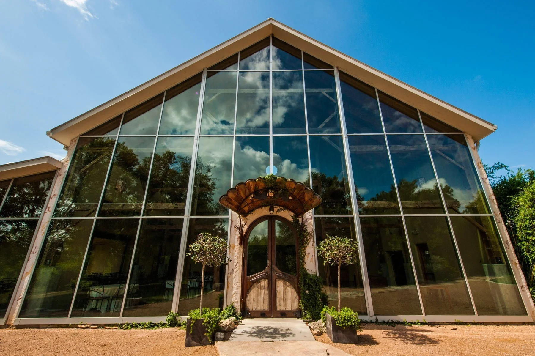 A modern building with large glass windows reflecting the blue sky and clouds, featuring a peaked roof and a decorative archway entrance surrounded by small trees and greenery.
