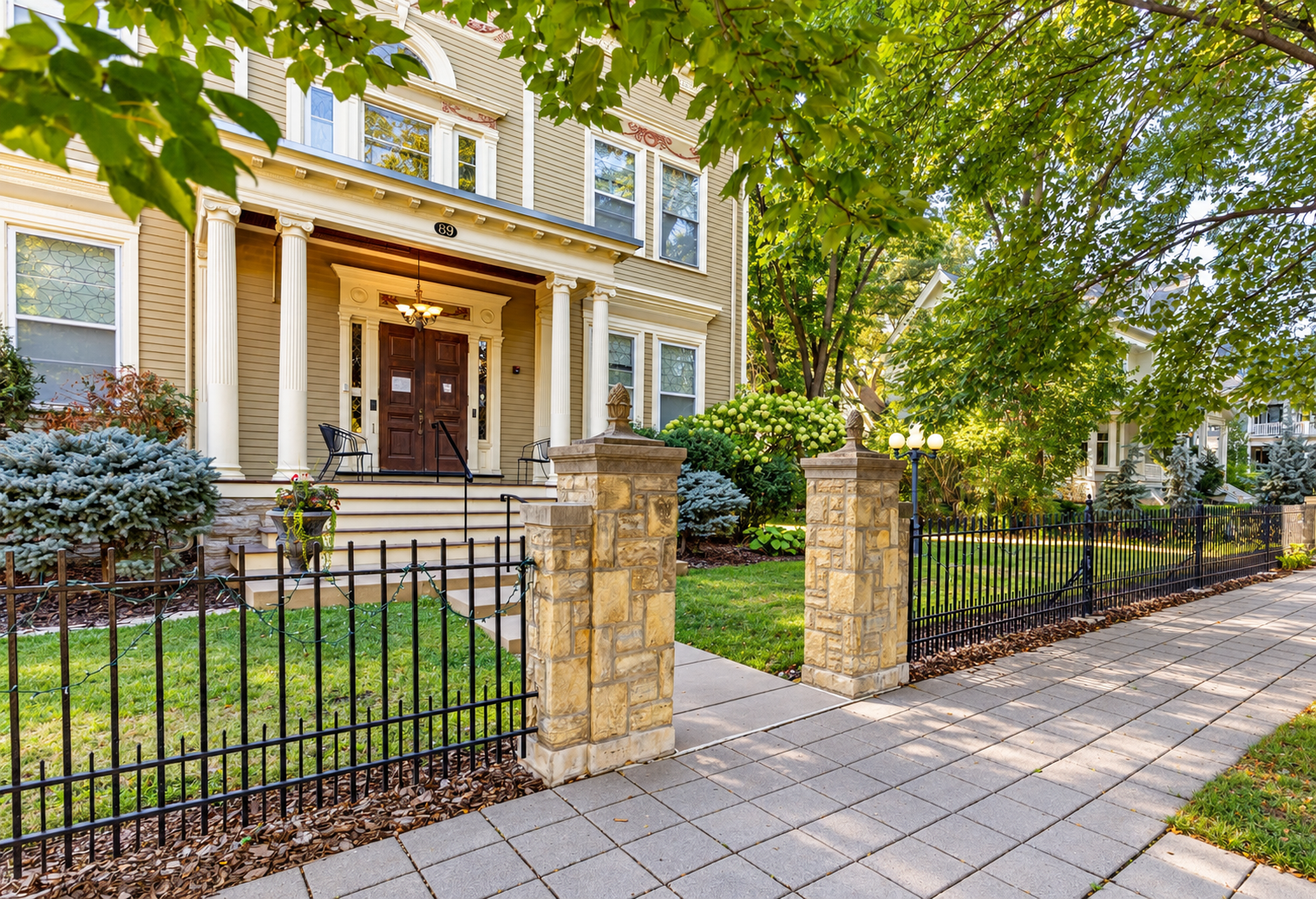 Front yard of a house with a stone and black metal fence, steps leading to a wooden front door, surrounded by trees and green shrubs under sunlight.