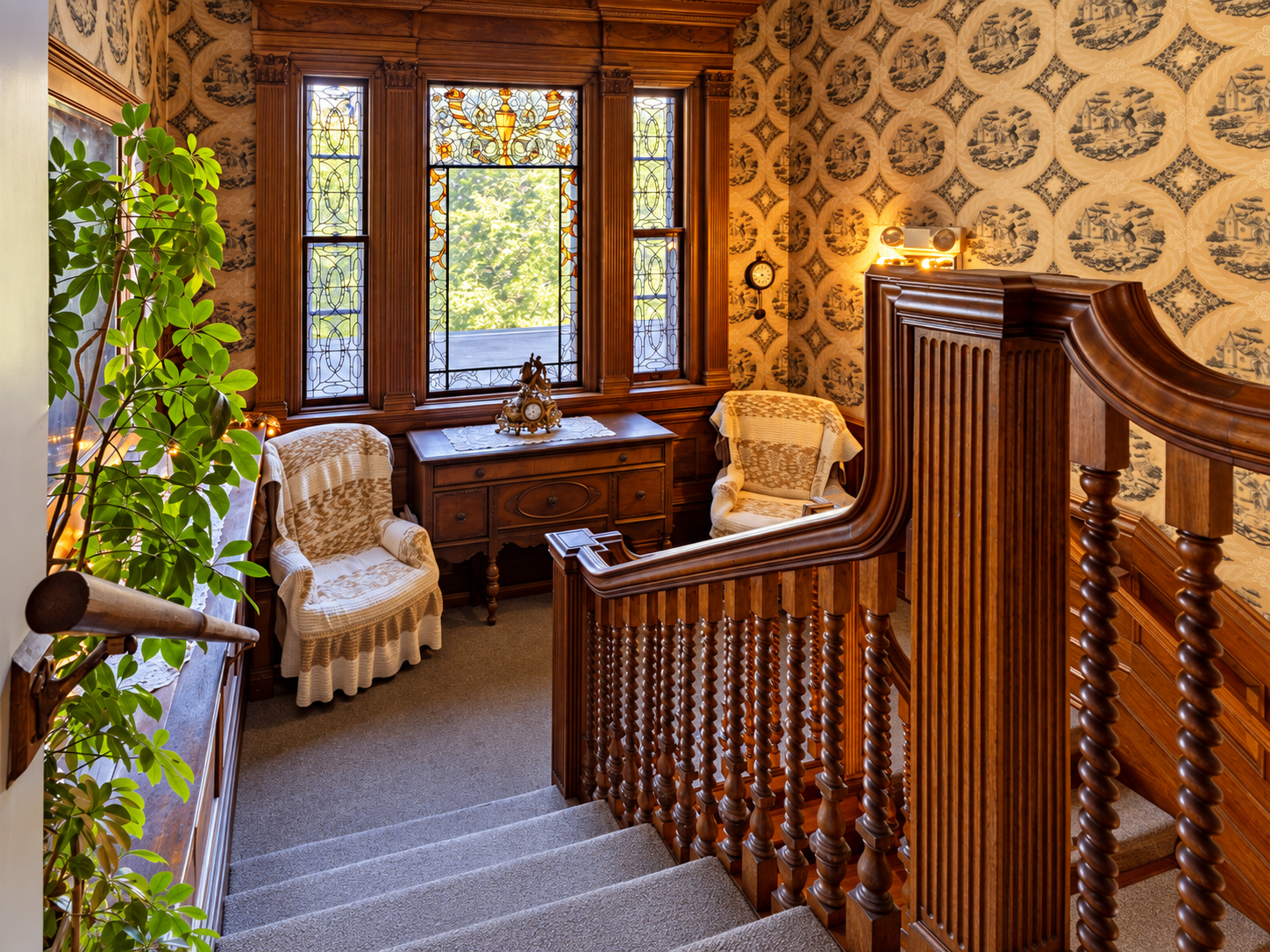 A cozy, vintage-style sitting area with wood accents, two armchairs with lace covers, a side table, decorative wallpaper, a stained glass window, a large leafy plant, and a carpeted staircase.