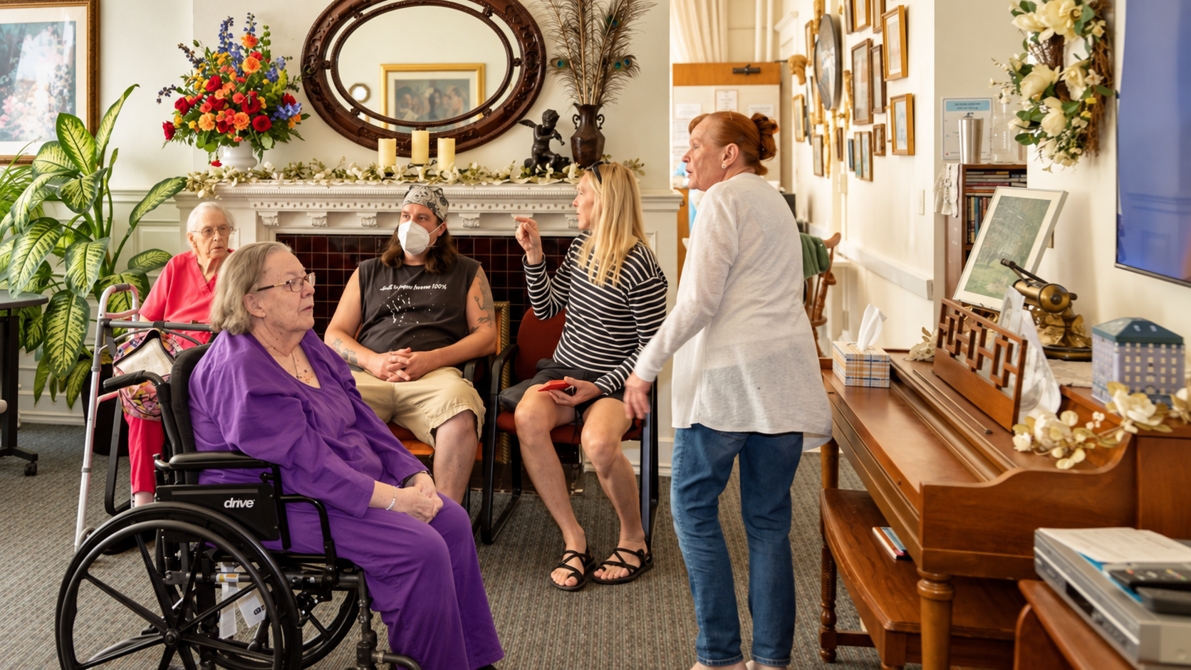 A group of elderly women and a caregiver are having a conversation in a cozy, well-decorated room with a fireplace, plants, framed pictures, and a piano.