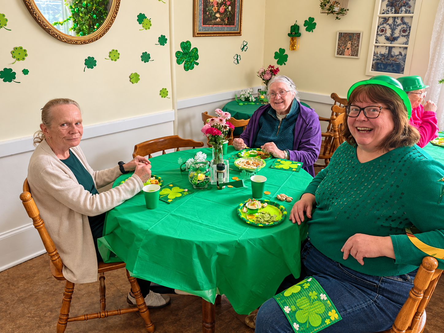 Three women celebrating St. Patrick's Day indoors, seated around a table decorated with green tablecloths, shamrocks, flowers, and St. Patrick's Day-themed tableware. They are wearing green accessories and smiling.