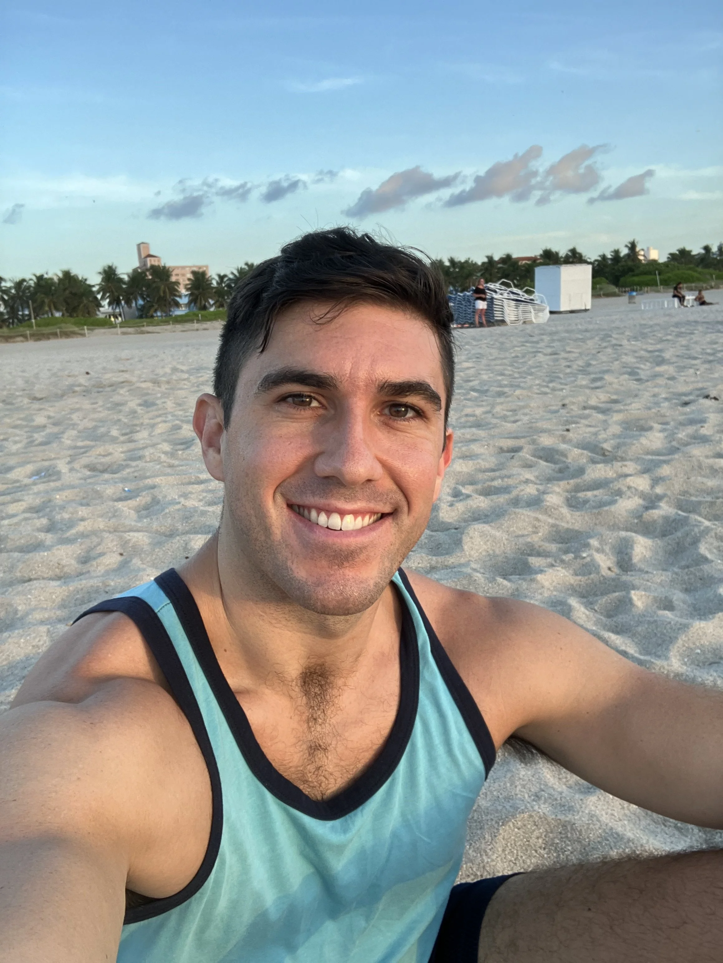 Zoomed in selfie of a smiling young man with short dark hair on a beach, wearing a blue sleeveless shirt, with sand and some beach structures and trees in the background.