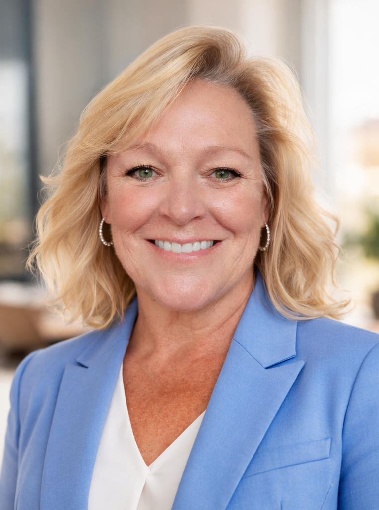 A smiling middle-aged woman with blonde hair, wearing a blue blazer and pearl earrings, in an office setting.