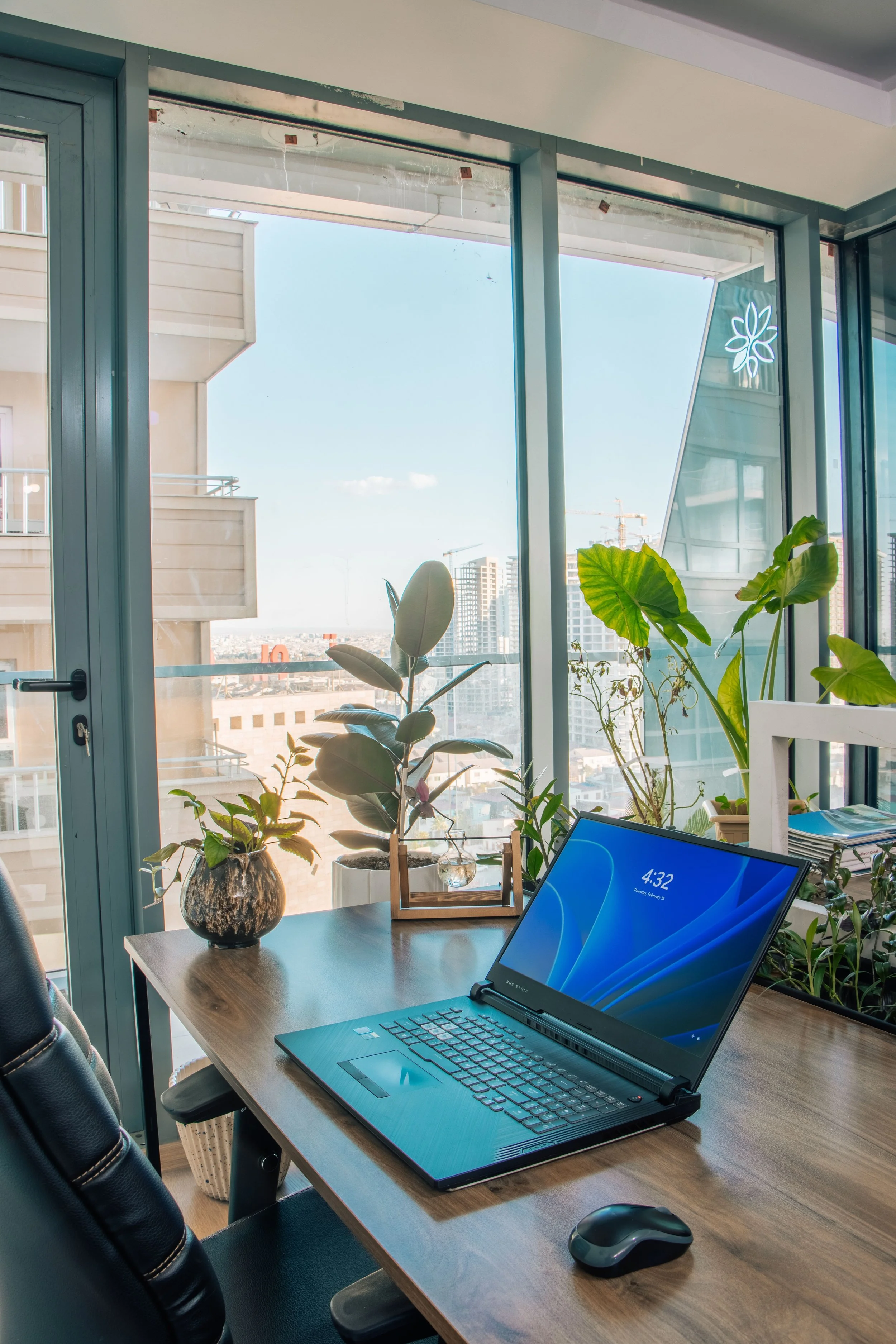 Interior of an office with a wooden desk, a laptop, a computer mouse, and several potted plants near a large window showing a cityscape with buildings and a construction crane in the distance.