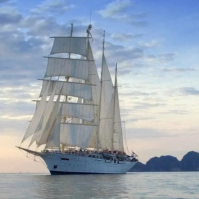 Segelschiff mit weißen Segeln auf ruhigem Wasser bei Sonnenuntergang, im Hintergrund eine Insel.