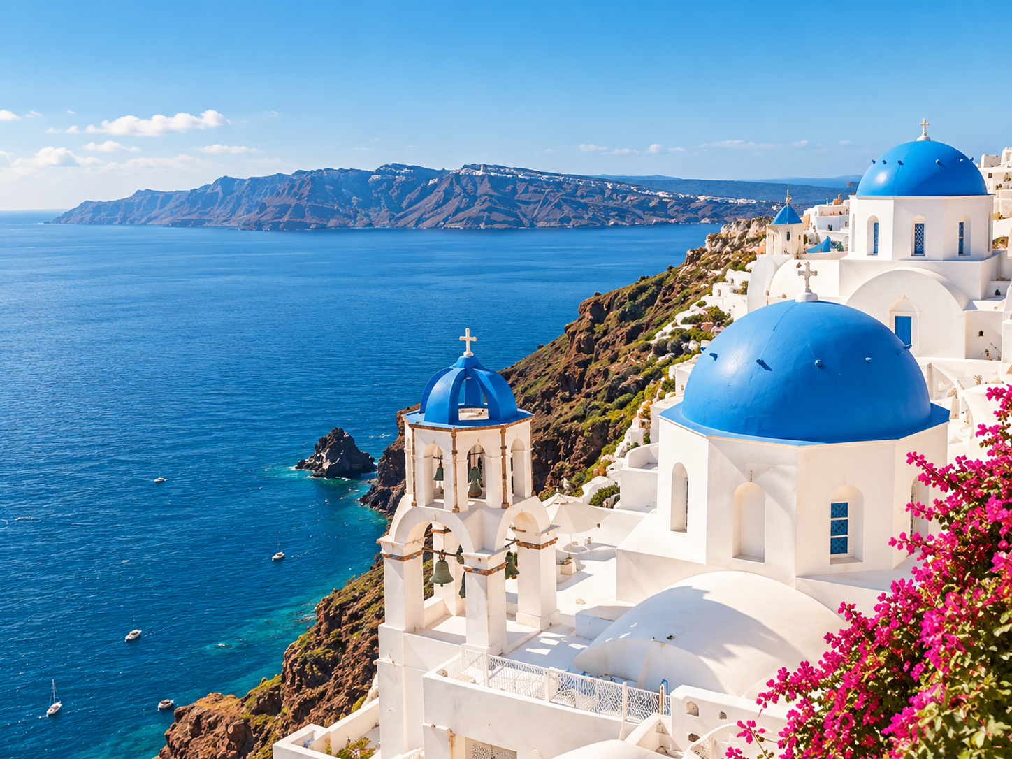 Weiße Kirchen mit blauen Kuppeln auf einer Steilküste in Santorini, Griechenland, mit Blick auf das Ägäische Meer und eine Bergkette im Hintergrund.