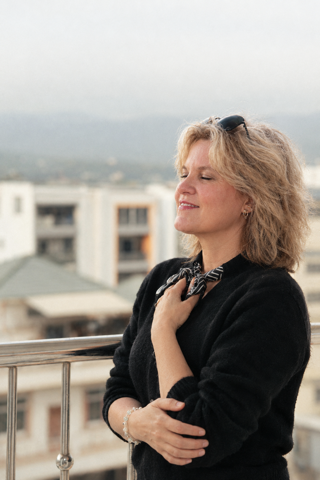 Eine Frau mit blonden, lockigen Haaren steht auf einem Balkon, trägt eine schwarze Kleidung, eine schwarze Schleife am Hals und eine Brille auf dem Kopf, und genießt die Aussicht bei Sonnenuntergang.