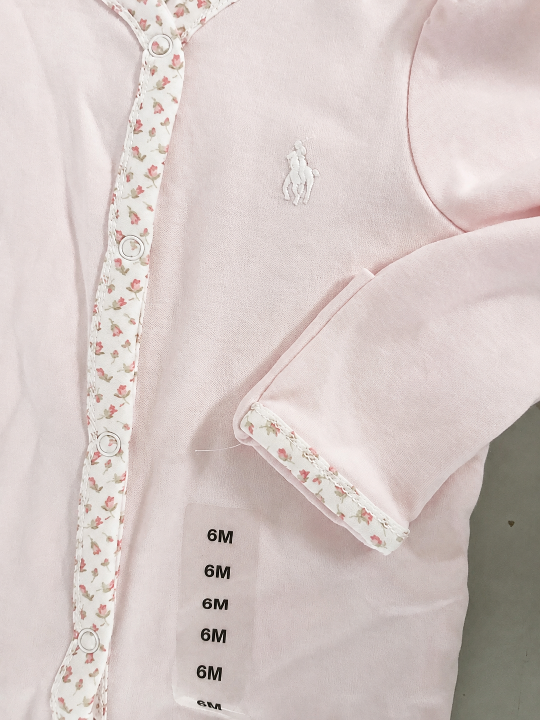 Close-up of a light pink baby onesie with floral trim, branded with the Polo Ralph Lauren logo, and a size tag showing 6 months.