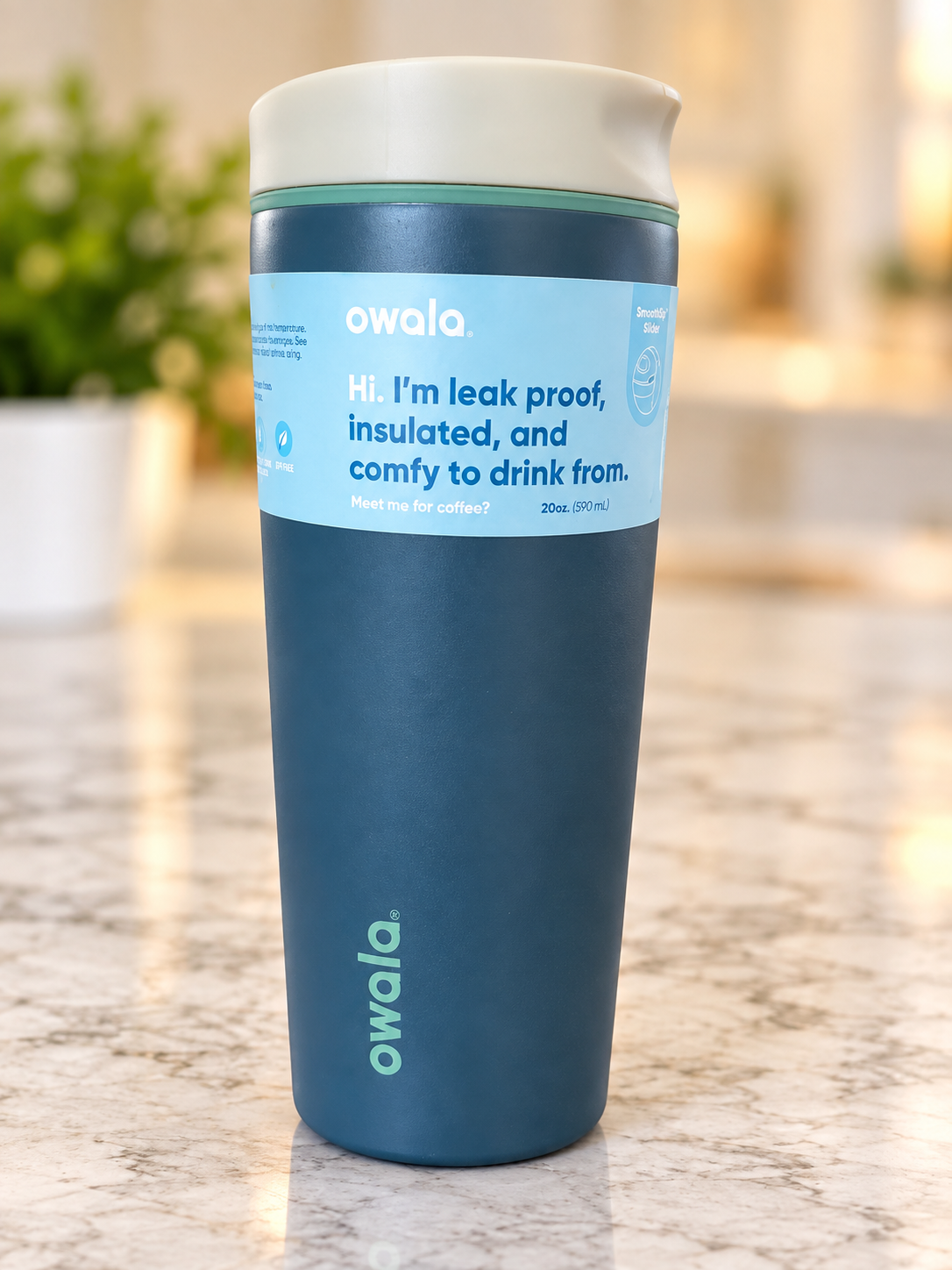 A blue insulated travel mug with a white lid, featuring a label that reads 'Hi. I'm leak proof, insulated, and comfy to drink from,' on a marble countertop.