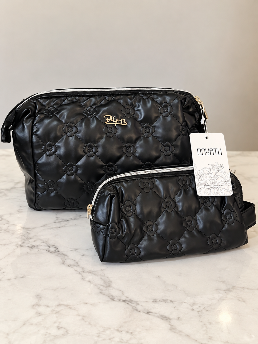 Two black quilted bags with floral patterns on a marble surface, one large and one small, both with gold accents and tags attached.