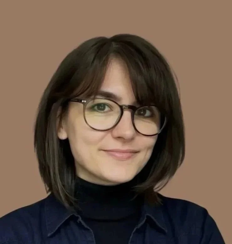 A woman with short brown hair, glasses, and a black turtleneck under a dark blue shirt, smiling against a plain brown background.