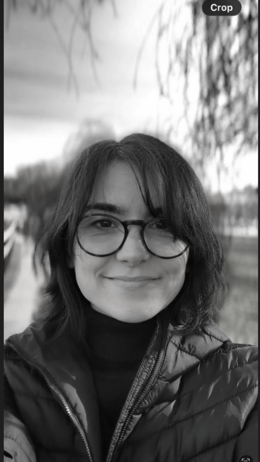 A smiling person with shoulder-length hair and glasses, wearing a jacket, outdoors near a body of water with trees in the background.