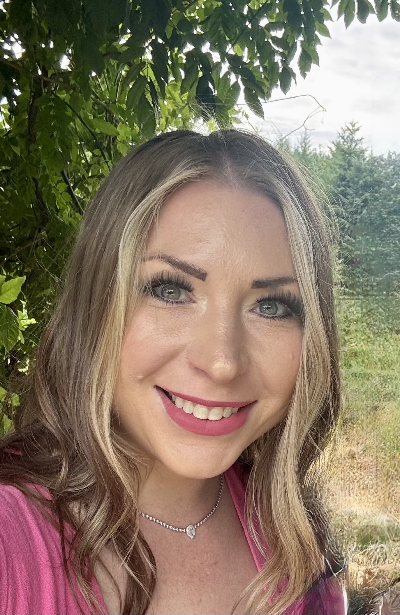 A woman with blonde hair, makeup, and a pink top smiling outdoors among green foliage and trees.