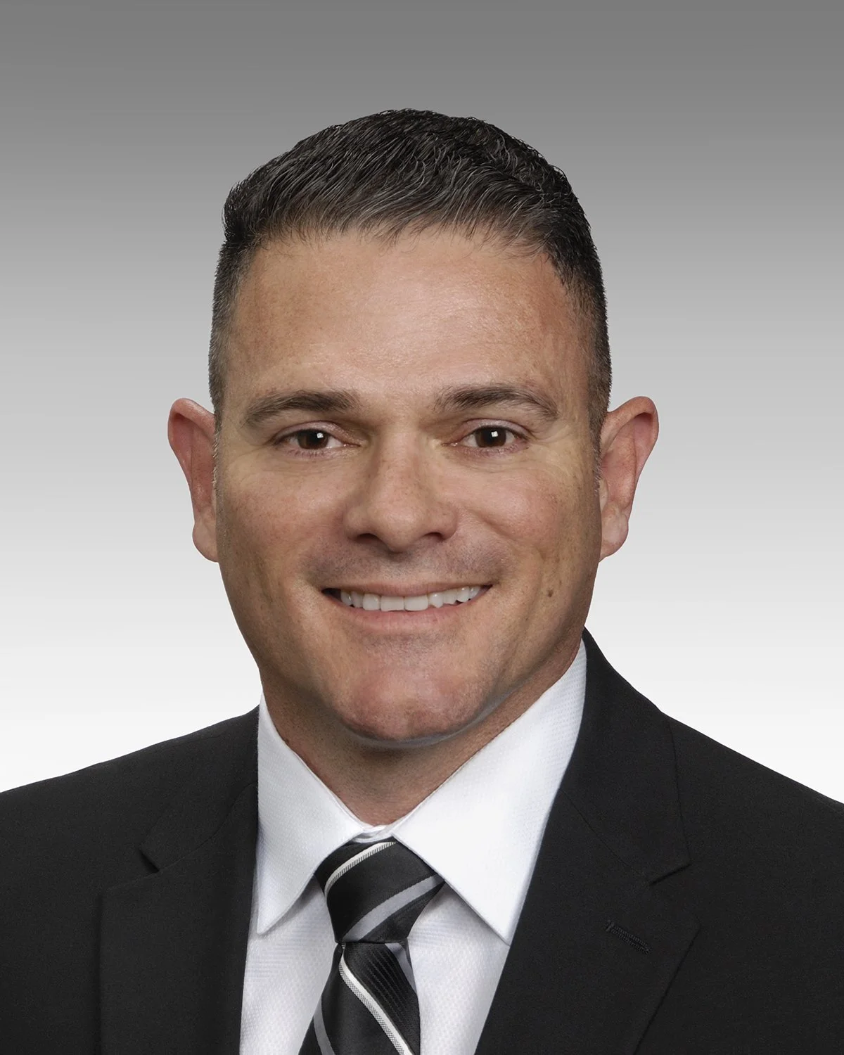 A professional headshot of a smiling man in a suit with a white shirt and striped tie, against a gradient gray background.