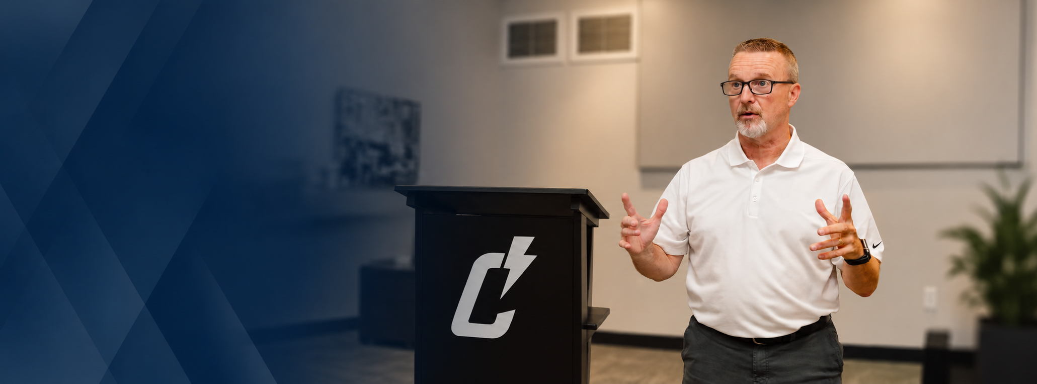 A man with glasses and a white polo shirt giving a presentation behind a black podium with a lightning bolt logo in a modern conference room.