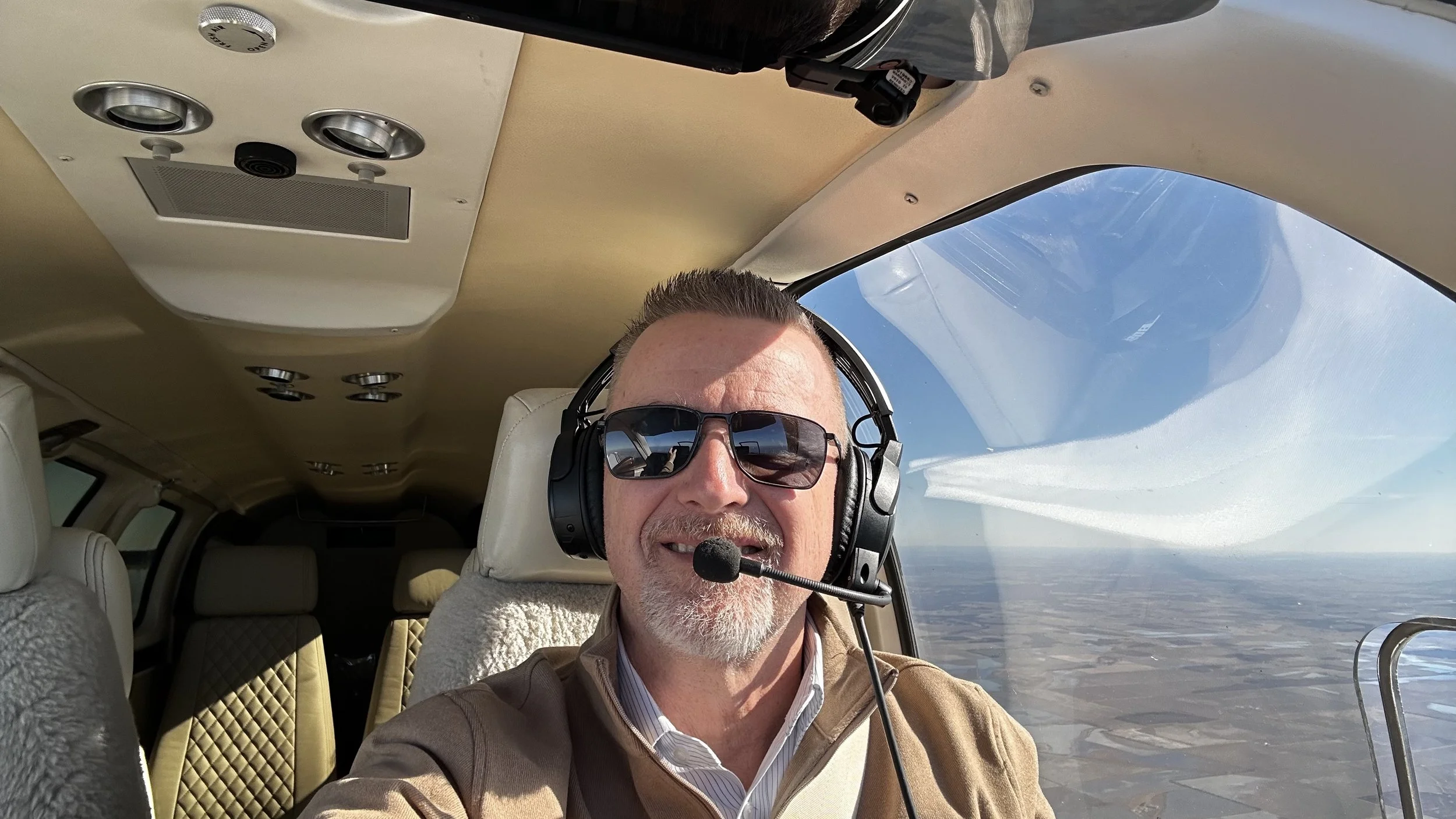A man with sunglasses, a beard, and a headset is flying in a small plane, with a view of the sky and landscape below through the window.